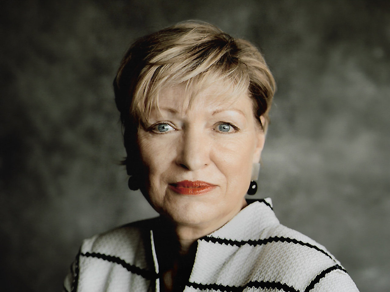 Karin Bergmann mit kurzem, blondem Haar blickt direkt in die Kamera. Sie trägt eine helle Jacke mit schwarzen Linien und auffällige, dunkle Ohrringe. Der Hintergrund ist dunkel und wirkt strukturiert. Ihr Gesichtsausdruck ist neutral und ruhig.