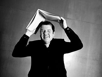 Ivor Bolton steht vor einer glatten Wand und hält ein geöffnetes Notenbuch über seinem Kopf, sodass die Seiten wie ein Dach wirken. Er trägt einen dunklen Anzug und lächelt direkt in die Kamera. Die Aufnahme ist schwarz-weiß und zeigt eine Nahaufnahme, die den Oberkörper und das Gesicht betont.