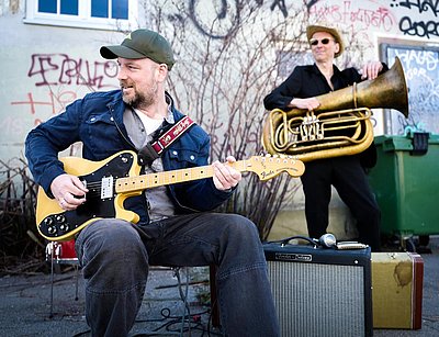 Stephan Zinner sitzt im Vordergrund auf einem Stuhl und spielt eine E-Gitarre. Er trägt eine blaue Jeansjacke, ein helles T-Shirt und eine Baseballkappe. Die Gitarre ist mit einem auffälligen Gurt versehen. Neben ihm steht ein Gitarrenverstärker. Im Hintergrund steht eine weitere Person mit einer Tuba, beide befinden sich im Freien vor einer Wand mit Graffiti und kahlen Ästen.