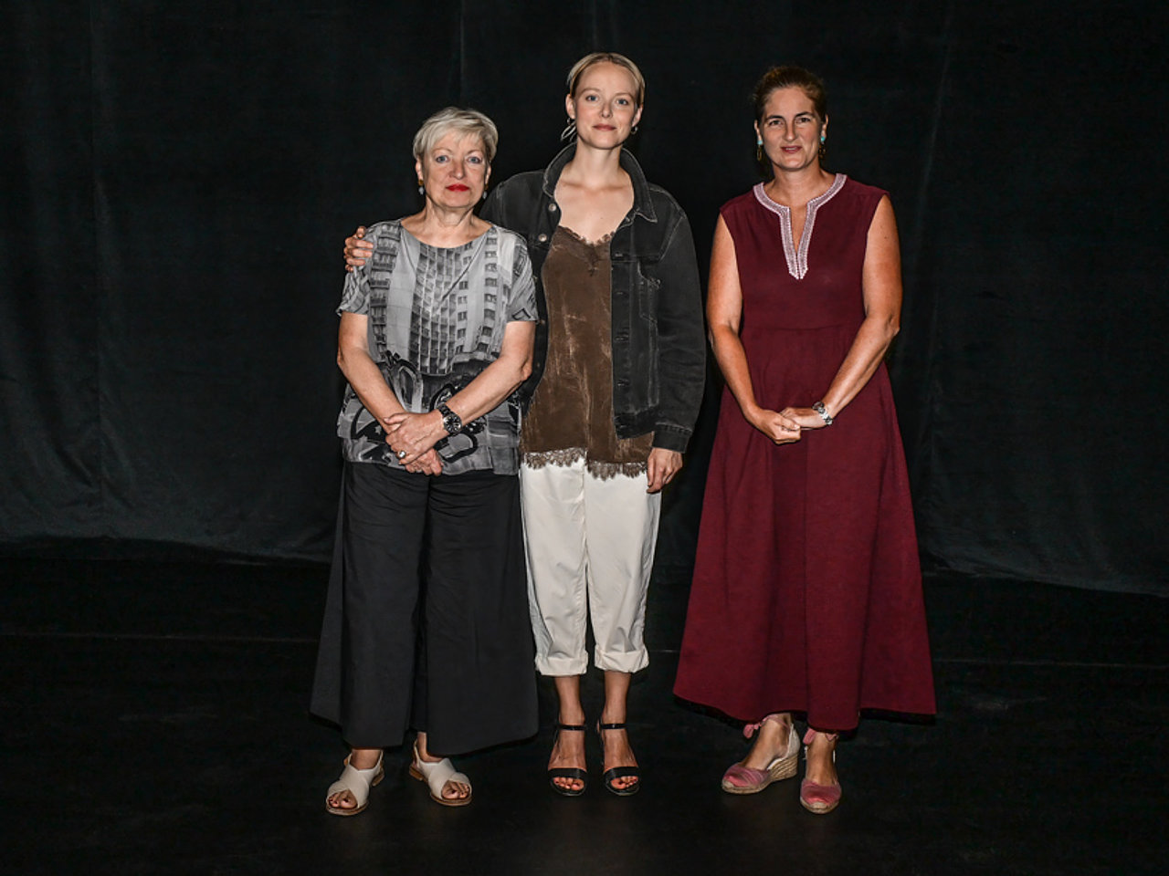 Karin Bergmann, Marie Luise Stockinger und Johanna Mitterbauer stehen nebeneinander vor einem dunklen Hintergrund. Karin Bergmann trägt eine gemusterte Bluse, eine dunkle Hose und rote Lippen. Marie Luise Stockinger trägt eine helle Hose, ein braunes Oberteil und eine dunkle Jacke, dazu offene Sandalen. Johanna Mitterbauer trägt ein ärmelloses, bodenlanges Kleid in dunklem Rot und hat die Hände vor dem Körper verschränkt. Alle blicken direkt in die Kamera.