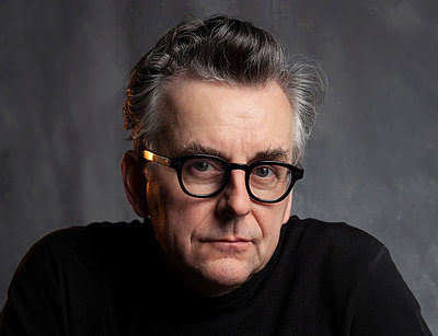 Johannes Silberschneider blickt direkt in die Kamera. Er trägt eine runde, dunkle Brille mit goldfarbenen Bügelenden und ein schwarzes Oberteil. Das Gesicht ist neutral, die Haare sind grau und sorgfältig gestylt. Der Hintergrund ist einfarbig und dezent, wodurch das Gesicht im Vordergrund steht.
