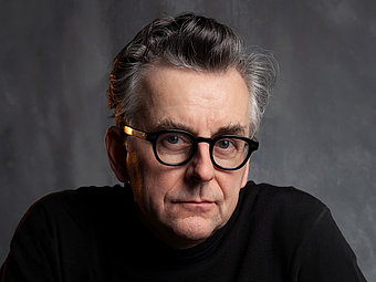 Johannes Silberschneider blickt direkt in die Kamera. Er trägt eine runde, dunkle Brille mit goldfarbenen Bügelenden und ein schwarzes Oberteil. Das Gesicht ist neutral, die Haare sind grau und sorgfältig gestylt. Der Hintergrund ist einfarbig und dezent, wodurch das Gesicht im Vordergrund steht.