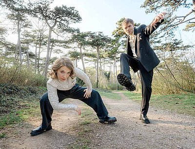 Zwei Mitglieder von Wiener Blond posieren auf einem Waldweg in ausdrucksstarker Haltung, beide tragen elegante Kleidung und schauen direkt in die Kamera.